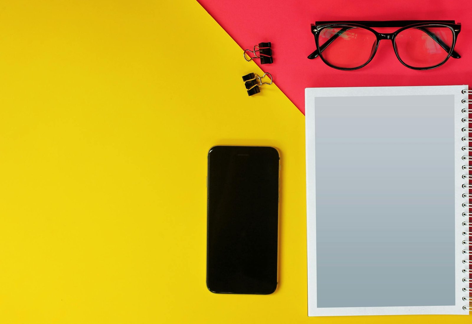 Modern flat lay featuring smartphone, glasses, notebook on vibrant yellow and red background.